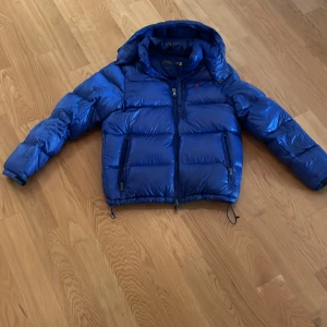 Blå pufferjacka från Polo Ralph Lauren - Snygg blå pufferjacka från Polo Ralph Lauren med glansig finish och huva. Jackan har dragkedja framtill, två stora fickor med dragkedja och justerbara snören i nederkant. Perfekt för kalla dagar och riktigt skön att bära.