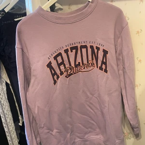 Ljuslila sweatshirt Arizona H&M  - Säljer en ljuslila sweatshirt från H&M i storlek M med tryck 'Arizona Phoenix' på bröstet. Tröjan har rund hals, långa ärmar och en relaxed fit. Perfekt för dig som gillar oversize tröjor och vill ha något bekvämt och snyggt.