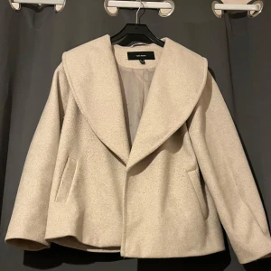 Beige kappa från Vero Moda - Stilren beige kappa från Vero Moda med bred krage och rak passform. Jackan har två fickor framtill och är helfodrad. Perfekt för dig som gillar en clean och modern look med lite oversized känsla.