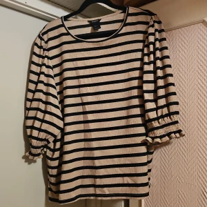 Randig topp från Lindex i storlek L - Beige och svart randig topp från Lindex med trekvartsärm och rynkade muddar. Tillverkad i mjuk polyesterblandning med lite stretch. Perfekt för dig som gillar en avslappnad men ändå snygg stil.