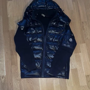 Moncler cardigan Navy Blue intressekoll! - Snygg svart pufferjacka från Moncler med glansig framsida och huva. Jackan har stickade ärmar och Moncler-logga på ena ärmen. Dragkedja framtill och fickor med tryckknappar. Perfekt för dig som vill ha en stilren och sportig look. Sitter perfekt på ca 1,83. Finns en del defekter så kom privat för flera bilder!