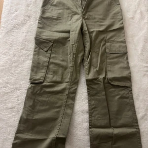 Olivgröna cargopants med stora fickor - Säljer ett par olivgröna cargopants med bred passform och stora fickor på sidorna. Byxorna har klassiska cargodetaljer och är tillverkade i bomull, vilket gör dem både bekväma och slitstarka. Perfekta för en avslappnad och trendig stil.
