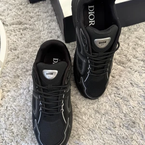 Svarta Dior B30 sneakers - Säljer ett par svarta Dior B30 sneakers med coola reflekterande detaljer och tydlig Dior-logga på plösen. Skorna har en sportig siluett, snörning och är tillverkade i mesh och syntetmaterial för en modern vibe. Perfekta för dig som vill sticka ut med lyxig streetstyle. Med sko lådan 