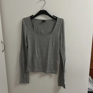 Grå långärmad topp med rund hals - Enkel och stilren grå långärmad topp från Gina Tricot med rundad, djup halsringning. Tillverkad i mjuk bomullsmix som känns skön mot huden. Perfekt att matcha med jeans eller kjol för en clean look.