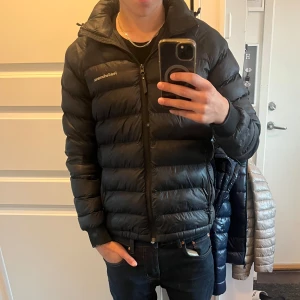 Svart dunjacka från Peak Performance - Svart dunjacka från Peak Performance med huva och dragkedja framtill. Perfekt för kalla dagar och har en clean, sportig look. Klassisk passform och diskret logga på bröstet. Den är tyvärr lite trasig på armbågen (se bild)