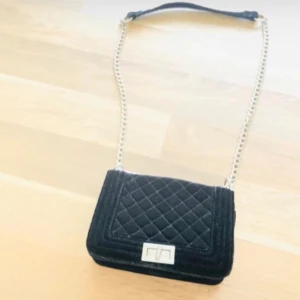 Svart quiltad axelväska med kedja - Snygg svart axelväska i sammet med quiltat mönster och silverfärgad kedjerem. Väskan har en stilren fyrkantig form och ett elegant spänne framtill. Perfekt för dig som vill ha en trendig och klassisk accessoar. Jätte fin och har används men väldigt vacker fortfarande. Kolla in mina andra väskor!