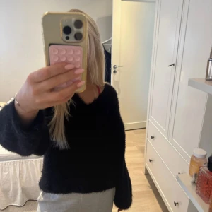 Svart fluffig stickad tröja Gina Tricot - Mysig svart stickad tröja från Gina Tricot med fluffig yta och v-ringad hals. Tröjan har långa ärmar och en loose passform som ger en chill vibe. Perfekt för dig som gillar mjuka och sköna plagg med lite extra volym i ärmarna.