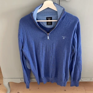 Blå GANT tröja med half zip - Blå tröja från GANT i 100% bomull med halv dragkedja och broderad logga på bröstet. Långärmad modell med ribbade muddar och ljusare armbågslappar som snygg detalj. Perfekt för dig som gillar klassisk och stilren look. Det står att den är large men den passar mer som en M/S