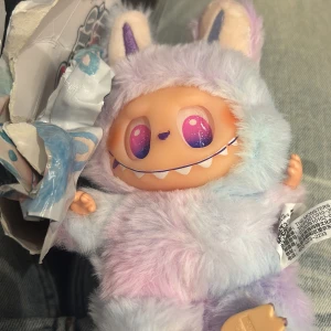 Man får den i boxen, men är öppnad ❤️ - Detta är en söt samlarfigur i pastellfärgad plyschdräkt med stora, glittrande ögon och kaninöron. Figuren har ett charmigt leende och mjuk päls i lila, blått och rosa. Förpackningen är skadad men själva figuren ser ut att vara i mycket gott skick utan synliga defekter. En labubu 