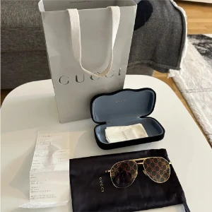 Gucci aviator solglasögon med GG-mönster - Snygga aviator solglasögon från Gucci med guldfärgade metallbågar och GG-logga i orange och guld över hela linsen. Glasen är tonade och har en unik design som verkligen sticker ut. Kommer med originalfodral, putsduk och Gucci-påse.