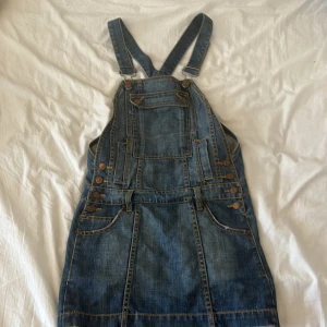 Jeansklänning med hängslen från FCUK - Säljer en cool jeansklänning från FCUK Jeans i klassisk blå denim. Klänningen har justerbara hängslen, fickor fram och bak samt snygga bronsfärgade knappar på sidorna. Perfekt för dig som gillar streetstyle och vill ha en avslappnad look.