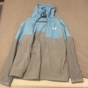 Blå och grå vindjacka från Under Armour - Snygg tvåfärgad vindjacka från Under Armour i ljusblått och grått. Jackan har huva, dragkedja framtill, lång ärm och logga på bröstet. Perfekt för blåsiga dagar och sportiga outfits. Materialet är lätt och känns smidigt mot kroppen.