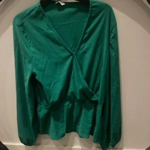 Grön omlottblus med resårärm - Snygg grön omlottblus från C i storlek M. Blusen har långa ärmar med resår vid ärmslut och en markerad midja. Materialet är lätt och har en diskret struktur som ger en cool känsla. Perfekt för dig som gillar en trendig och bekväm look.