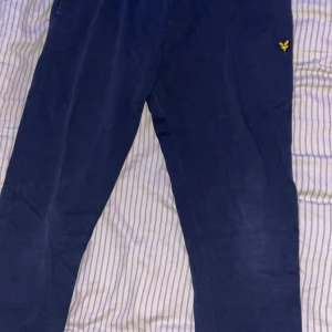 Blå mjukisbyxor från Lyle & Scott - Mörkblå mjukisbyxor från Lyle & Scott med klassisk gul logga broderad på ena benet. Byxorna har resår i midjan och en bakficka. Perfekta för chill eller träning, tillverkade i mjukt bomullsmaterial.