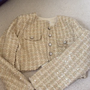 Beige kavajjacka med paljetter - Säljer en beige kavajjacka i rutig struktur med glittriga paljettdetaljer. Jackan är croppad, har långa ärmar, två fickor framtill och knäpps med knappar. Perfekt för dig som vill sticka ut lite extra med en chic och trendig look.