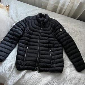 Svart dunjacka från Moncler - Svart dunjacka från Moncler med quiltad design och hög krage. Jackan har två fickor med dragkedja framtill och Moncler-logga på ärmen. Snygg och stilren modell med raka linjer och diskreta detaljer. Perfekt för kyliga dagar.