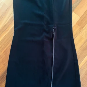 Svart långkjol med slits och knyt - Stilren svart långkjol från John Zack med rak passform och en snygg slits framtill som har ett knytband. Kjolen är enkel och elegant, perfekt för dig som gillar minimalistisk stil. Materialet känns mjukt och följsamt. 