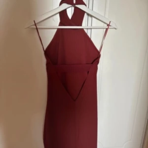 Vinröd halterneckklänning - Vinröd kortklänning med halterneck och smala axelband. Klänningen har en djup v-ringning i ryggen och är figurnära med stilren design. Perfekt för dig som vill sticka ut med en elegant och modern look.