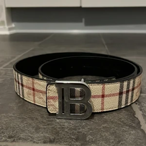 Burberry rutigt bälte med B-spänne - Snyggt bälte från Burberry med klassiskt rutigt mönster i beige, svart och rött. Bältet har ett stilrent metallspänne i form av ett stort B. Materialet är en mix av skinn och textil, perfekt för att lyfta din outfit med en lyxig touch.
