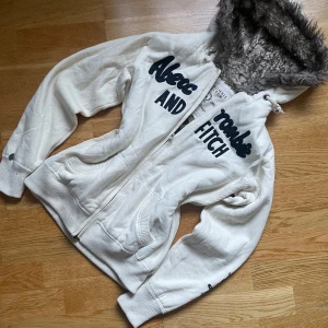 Abercrombie & Fitch y2K  zip up med päls  -  ❄️ Mysig Abercrombie & Fitch Hoodie (New York Edition) ❄️  ✨ Märke: Abercrombie & Fitch (New York Edition) 🗽 🎨 Färg: Vit 🧵 Material: Mjukt tyg med fuskpälsfodrad huva 👚 Design: Zip-up med svart text  Denna hoodie är perfekt för att hålla dig varm och stilfull under kyliga dagar! Fuskpälsen i huvan gör den extra bekväm och mysig. ☁️ Den vita färgen passar till allt och den exklusiva New York-versionen av Abercrombie & Fitch gör att du sticker ut från mängden. 🤩  