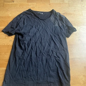 Svart t-shirt från Jack & Jones Core XL - Svart t-shirt från Jack & Jones Core i storlek XL. Klassisk modell med rund hals och korta ärmar. Tryck med små vita bokstäver på ena axeln och bröstet. Tillverkad i mjuk bomull för en skön känsla.