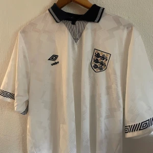England fotbollströja Umbro vit - Klassisk England fotbollströja från Umbro i vit med subtilt geometriskt mönster. Tröjan har svart krage med ränder, broderad landslagssköld och Umbro-logga på bröstet. Ärmarna är korta med svartvitt grafiskt band. Perfekt för dig som gillar retrostil.