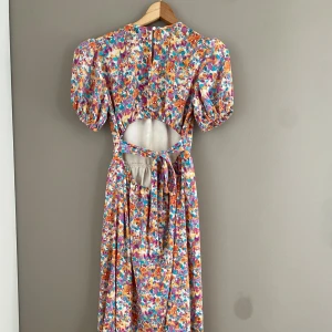 Blommig långklänning från Bik Bok - Jätte söt långklänning från Bik Bok med blommigt mönster i orange, lila, turkos och vitt. Klänningen har puffiga korta ärmar, rund halsringning, öppen rygg med knyt och lätt utställd kjol. Perfekt för dig som vill sticka ut med en lekfull och somrig stil.