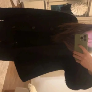 Svart kappa med knappar - Stilren svart kappa med klassisk rak passform och knäppning framtill. Jackan har långa ärmar och en enkel design som passar perfekt till höst och vinter. Materialet ser ut att vara ullblandning och kappan har en tidlös look.