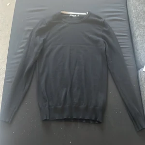 Svart stickad tröja från J.Lindeberg - Svart stickad tröja från J.Lindeberg med rund halsringning och diskret logga på bröstet. Tröjan har lång ärm och ribbade muddar vid ärmslut och nederkant. Perfekt för dig som gillar stilren och enkel design.