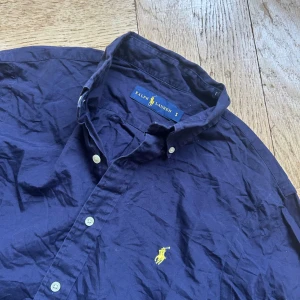 Mörkblå skjorta från Ralph Lauren - Klassisk mörkblå skjorta från Ralph Lauren med button-down krage och vita knappar. Ikoniska gula loggan broderad på bröstet. Skjortan är långärmad och tillverkad i mjuk bomull, perfekt för en clean och stilren look.