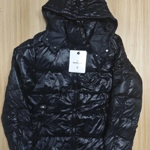 Svart dunjacka från Moncler med huva - Snygg svart dunjacka från Moncler med glansig finish och huva. Jackan har dragkedja framtill, två sidofickor med dragkedja och klassisk Moncler-logga på ärmen. Perfekt för kalla dagar och riktigt stilren look.