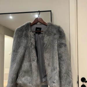 Grå pälsjacka från Bershka - Säljer en snygg grå pälsjacka från Bershka. Jackan har lång, fluffig fuskpäls och är öppen framtill utan synliga knappar eller dragkedja. Perfekt för dig som vill sticka ut med en trendig och mysig look.