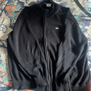 Svart stickad jacka från Lacoste - Svart stickad jacka från Lacoste med dragkedja framtill och ribbade muddar vid ärmslut och midja. Klassisk ståkrage med vit rand på insidan och den ikoniska gröna krokodilloggan på bröstet. Perfekt för dig som gillar stilren och sportig look.
