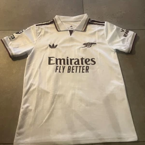 Arsenal vit fotbollströja Gyökeres S - Säljer en snygg Arsenal fotbollströja från Adidas i slim fit, storlek S. Tröjan är vit med mörkröda detaljer och har korta ärmar, krage och tryck med Emirates Fly Better på bröstet. På ryggen står Gyökeres och nummer 14. Materialet är lätt och andas.