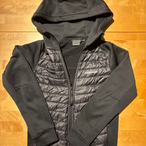 Svart hybridjacka från Didriksons - Svart hybridjacka från Didriksons med quiltad front och mjuka ärmar. Jackan har huva, dragkedja och diskret logga på bröstet. Perfekt för lager-på-lager och chill dagar. Materialmix med polyester och syntet, långärmad och normal passform. Liten skada som du ser på tredje bilden, därav lågt pris.