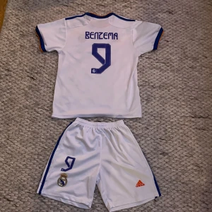 Real Madrid Benzema fotbollströja set - Snyggt Real Madrid matchställ med Benzema #9 på ryggen. Tröjan är vit med blå och orange detaljer, korta ärmar och klubbmärke på bröstet. Shorts i samma stil med nummer 9 och Real Madrid-logga. Tillverkad i lätt och ventilerande polyester. 