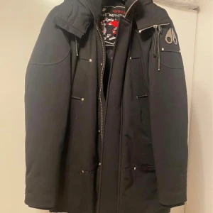 Svart dunjacka från Moose Knuckles - Svart dunjacka från Moose Knuckles med huva, silverfärgade detaljer och dragkedja. Jackan har flera fickor, broderad logga på axeln och ett coolt broderat innerfoder med hockeymotiv. Perfekt för kalla dagar och riktigt snygg streetstil.