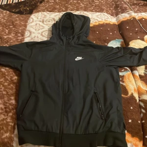 Svart Nike vindjacka med huva - Svart vindjacka från Nike med huva och dragkedja framtill. Jackan har två fickor med dragkedja och en vit Nike-logga på bröstet. Perfekt för en sportig och avslappnad stil.