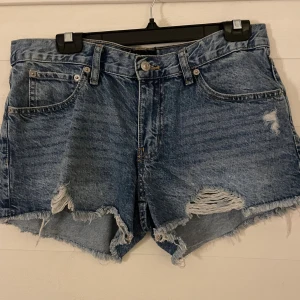 Blå jeansshorts med slitningar från 157 - Snygga blå jeansshorts från 157 med råa, fransiga kanter och slitna detaljer framtill. Klassisk femficksmodell med knapp och dragkedja. Perfekta för en avslappnad och trendig look i sommar.