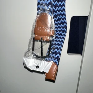 Blått zigzag-mönstrat bälte med skinn - Säljer ett snyggt bälte med blått och svart zigzag-mönster. Spännet är i metall och ändarna är förstärkta med brunt skinn. Perfekt för att ge outfiten lite extra edge och färg. Bältet är flexibelt och passar till jeans eller chinos.