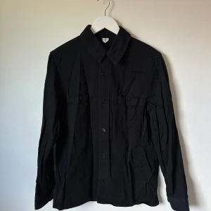 Svart overshirt med fickor, strl 48 - Svart overshirt med klassisk krage och två bröstfickor. Skjortan har knäppning framtill och långa ärmar med manschett. Perfekt att bära öppen eller stängd för en clean och stilren look. 3V13