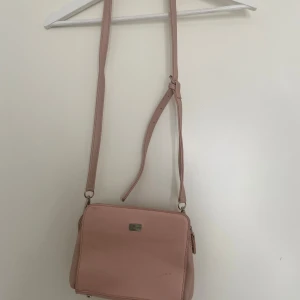 Rosa väska - Ljusrosa axelremsväska från David Jones Paris. Väskan har justerbar axelrem och silverfärgade detaljer. Den har två fack med dragkedja, även två mindre fack i de stora facken