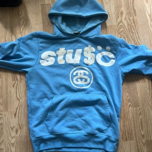 Blå Stussy hoodie med tryck, M - Säljer en blå hoodie från Stussy i 100% bomull. Stor vit logga och grafiskt tryck framtill, samt stor 8-ball och text på ryggen. Klassisk känguruficka och huva. Perfekt för dig som gillar streetwear och vill sticka ut med en unik design.