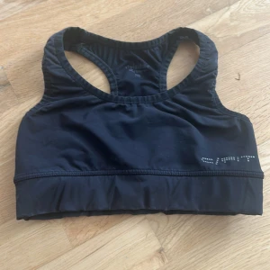 Svart sporttopp från SVEA - Sportig svart linnetopp från SVEA med brottarrygg och diskret logga framtill i silvriga detaljer. Tillverkad i stretchigt syntetmaterial som andas, perfekt för träning. Sitter tight och har bred resår nedtill för extra stöd.