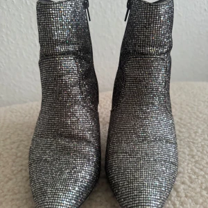 Glittriga silvriga stövletter med klack - Säljer ett par riktigt snygga stövletter i glittrigt silver med rutigt mönster. Skorna har spetsig tå, smal klack och dragkedja på sidan. Perfekta för dig som vill sticka ut och addera lite extra bling till outfiten. Använd en gång. 