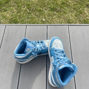 Nike Air Jordan 1 Mid Blå/Vit - Säljer ett par Nike Air Jordan 1 Mid i blått och vitt med snygga denimdetaljer och klassisk vit swoosh. Skorna har snörning, hög siluett och en cool mix av skinn och denimtyg. Perfekta för dig som vill sticka ut med en ikonisk sneaker. Size 40