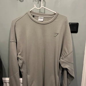 Oversized långärmad tröja från Gymshark - Säljer en stilren grå långärmad tröja från Gymshark i storlek S. Tröjan har en clean look med diskret logga framtill och stor Gymshark-tryck på ryggen. Perfekt för träning eller chill dagar. Tillverkad i mjukt material för maximal komfort. Perfekt pumpcover.