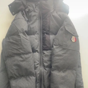 Grå Moncler pufferjacka  - Säljer en grå pufferjacka från Moncler med stor huva och dragkedja framtill. Jackan har en ficka med lock på ärmen och Moncler-logga. Fodrad och riktigt varm, perfekt för kalla dagar. Snygg och stilren design som sticker ut med sin puffiga look.