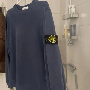 Blå sweatshirt från Stone Island - Säljer en blå sweatshirt från Stone Island med den klassiska loggan på vänster ärm. Tröjan har rund halsringning och långa ärmar. Tillverkad i mjuk bomull, perfekt för en avslappnad och stilren look.