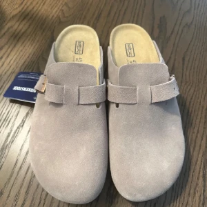 Beige mockasandaler från Birkenstock - Säljer ett par stilrena sandaler från Birkenstock i beige mocka. Modellen har en bred rem med metallknapp och präglad logga på sidan. Fotbädden är mjuk och anatomiskt utformad för extra komfort. Perfekta för en chill och avslappnad look.